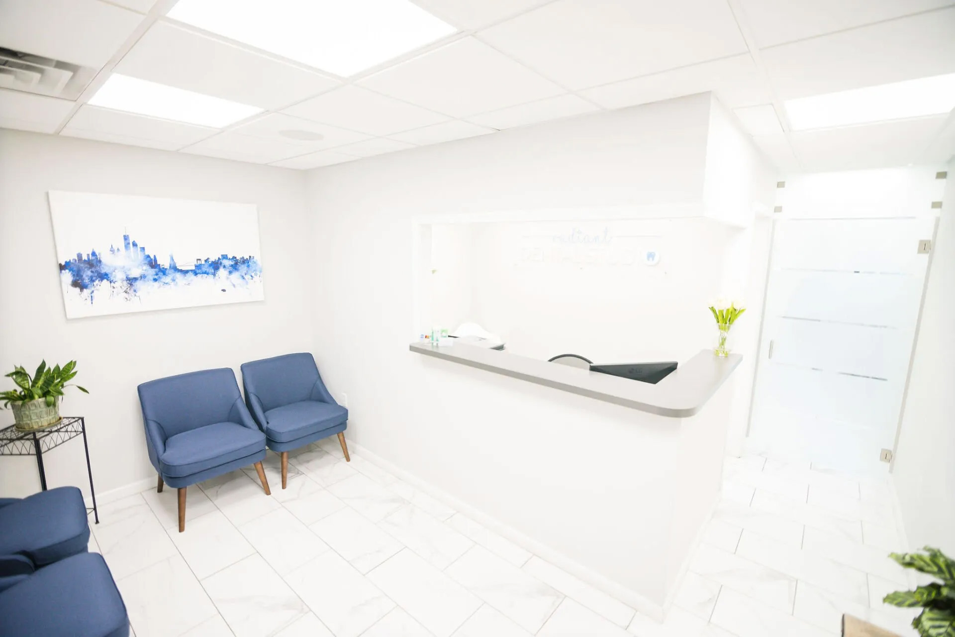 The image shows an interior space that appears to be a modern waiting room or reception area, featuring a clean and minimalist design with blue chairs, white walls, and a large window allowing natural light.