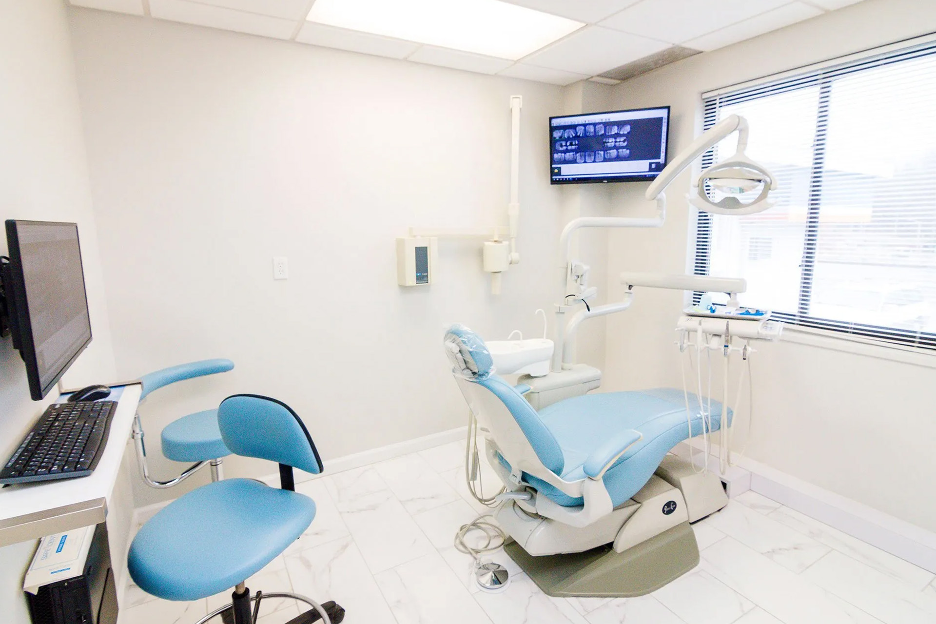 The image shows an interior view of a dental office with modern equipment, including a dental chair and a computer monitor displaying patient information.