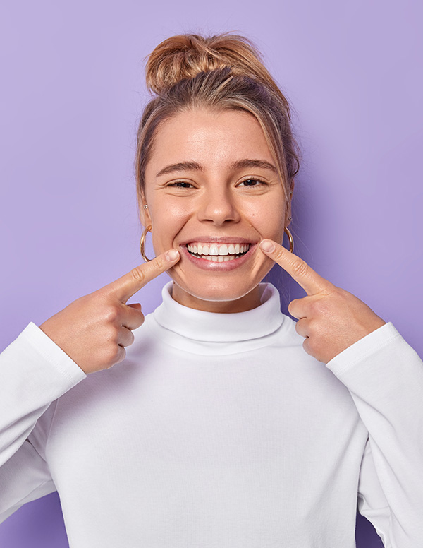 A woman is smiling broadly, holding her index finger to her lips in a playful manner.
