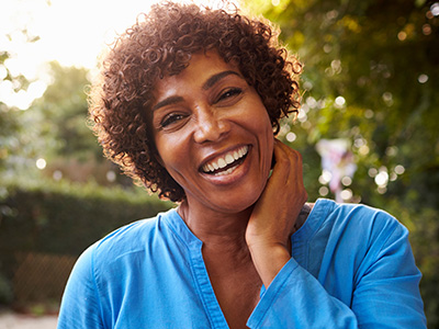 The image is of a woman with curly hair, smiling broadly at the camera. She has her hand on her chin and appears to be outdoors during daylight.