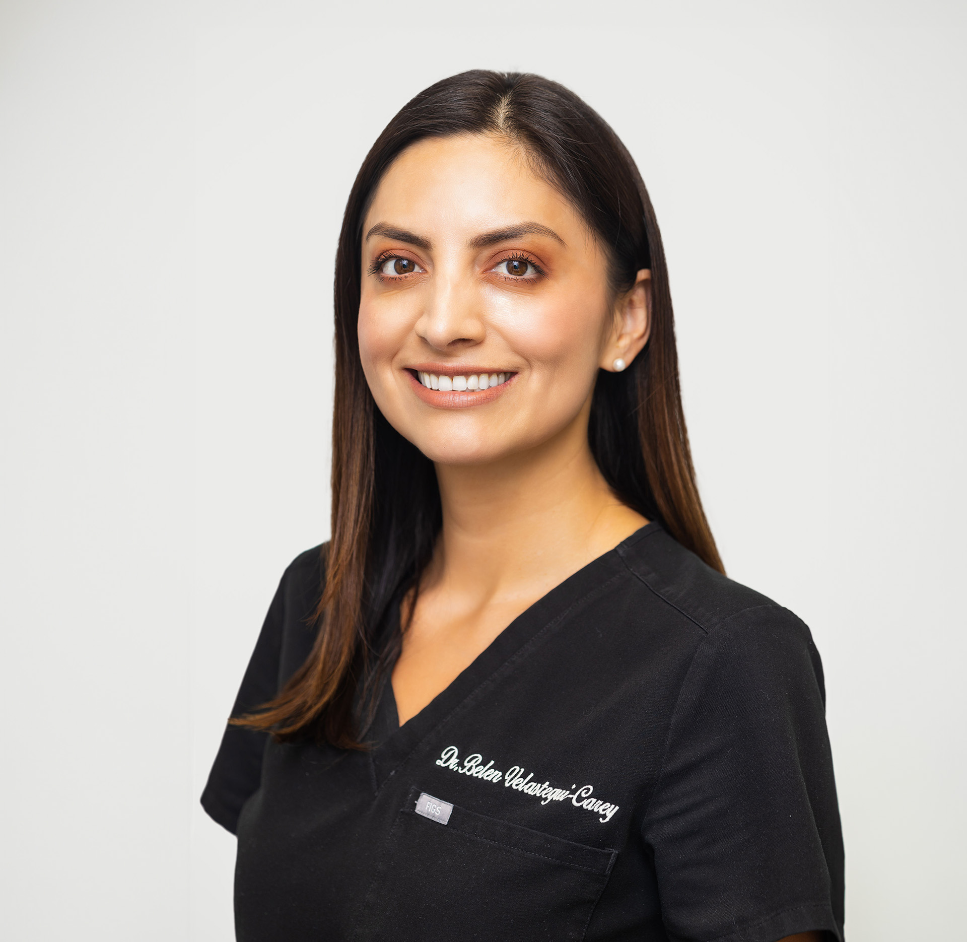 The image shows a woman wearing a white lab coat, standing in front of a dental office setup with a large screen displaying colorful graphics. She is smiling and appears to be posing for the photograph.