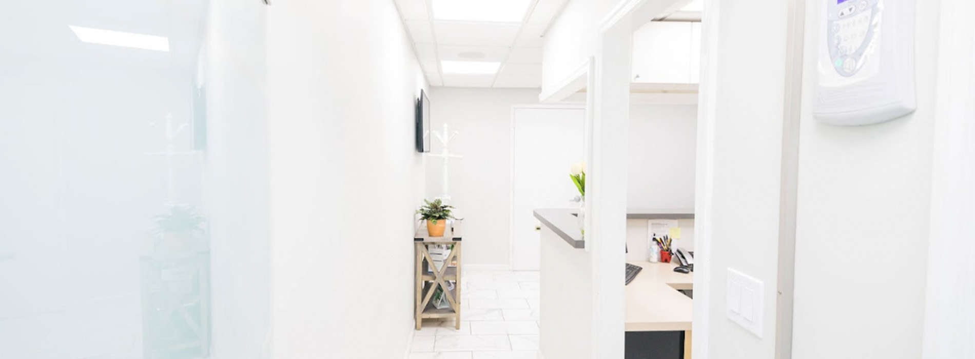 The image shows a modern interior space, likely an office or lobby, with a clean and minimalist design featuring white walls, glass partitions, a wooden floor, and a large window allowing natural light to enter the room.