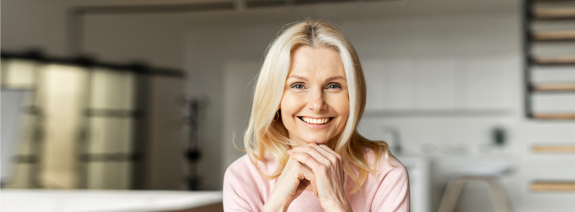 The image shows a woman with blonde hair, wearing a pink top, standing in a modern kitchen with white cabinets and appliances, looking directly at the camera.