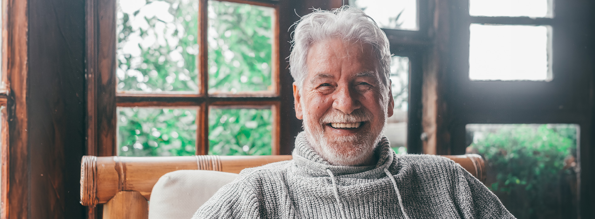 An elderly man with white hair and a beard, wearing a dark sweater, seated on a couch indoors.