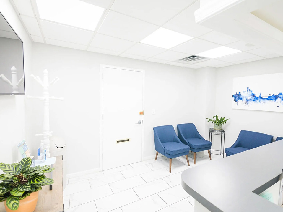 An interior view of a modern office space with a clean and minimalist design, featuring a reception area with seating, a desk with chairs, and a large window allowing natural light to enter.