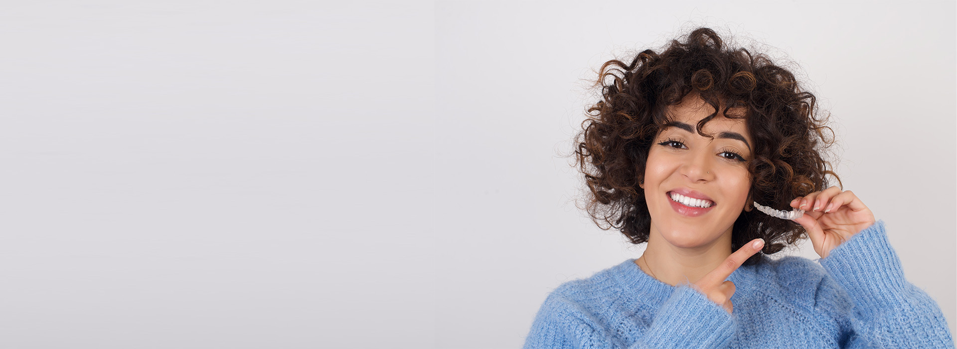 A smiling woman with curly hair, wearing a blue sweater, holding up her index finger.