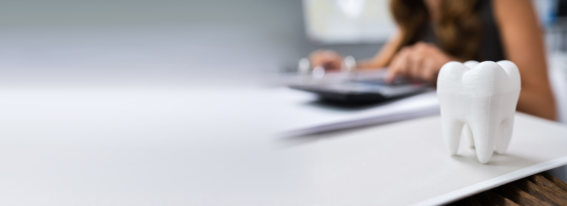 The image shows a person sitting at a desk with a laptop, working on papers or documents. In the foreground, there is a close-up of a toothbrush holder with a toothbrush inside. The background is blurred but appears to be an office setting.