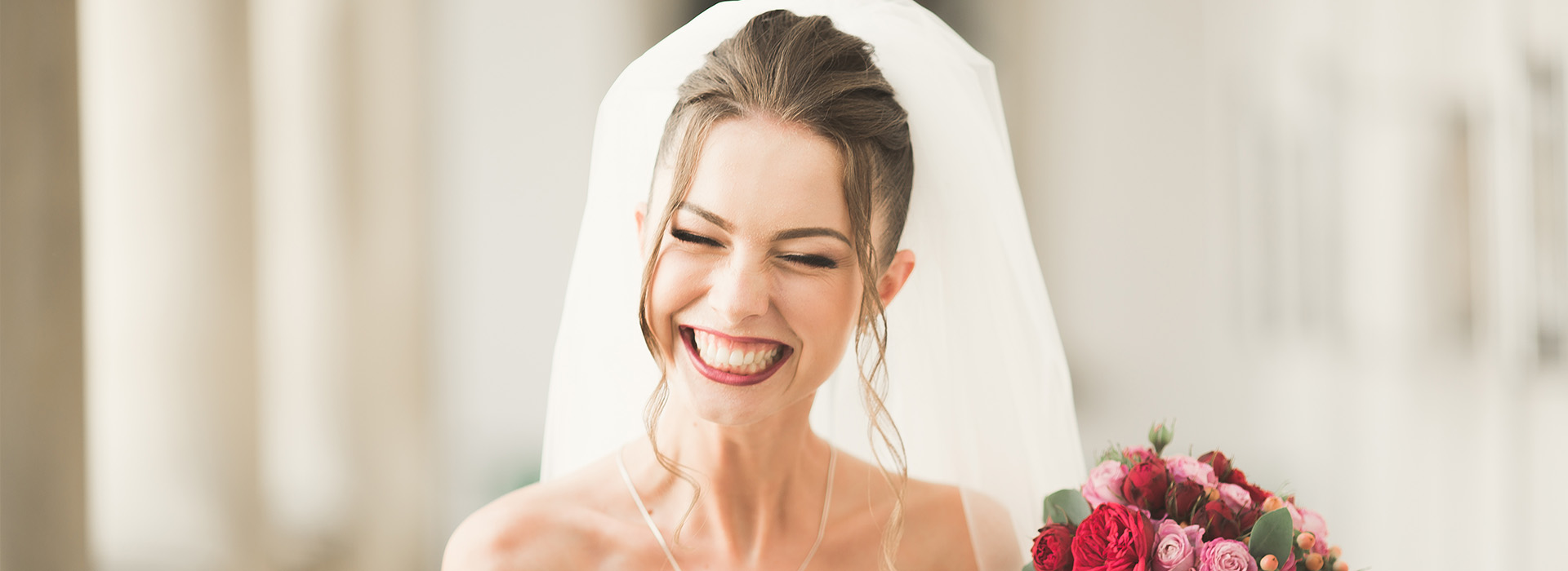 The image shows a bride wearing a white wedding dress and a necklace, standing beside a bouquet of red flowers, smiling at the camera.