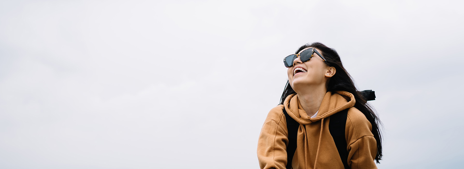 Woman in a brown jacket, sunglasses, and backpack smiles while standing against a clear sky.