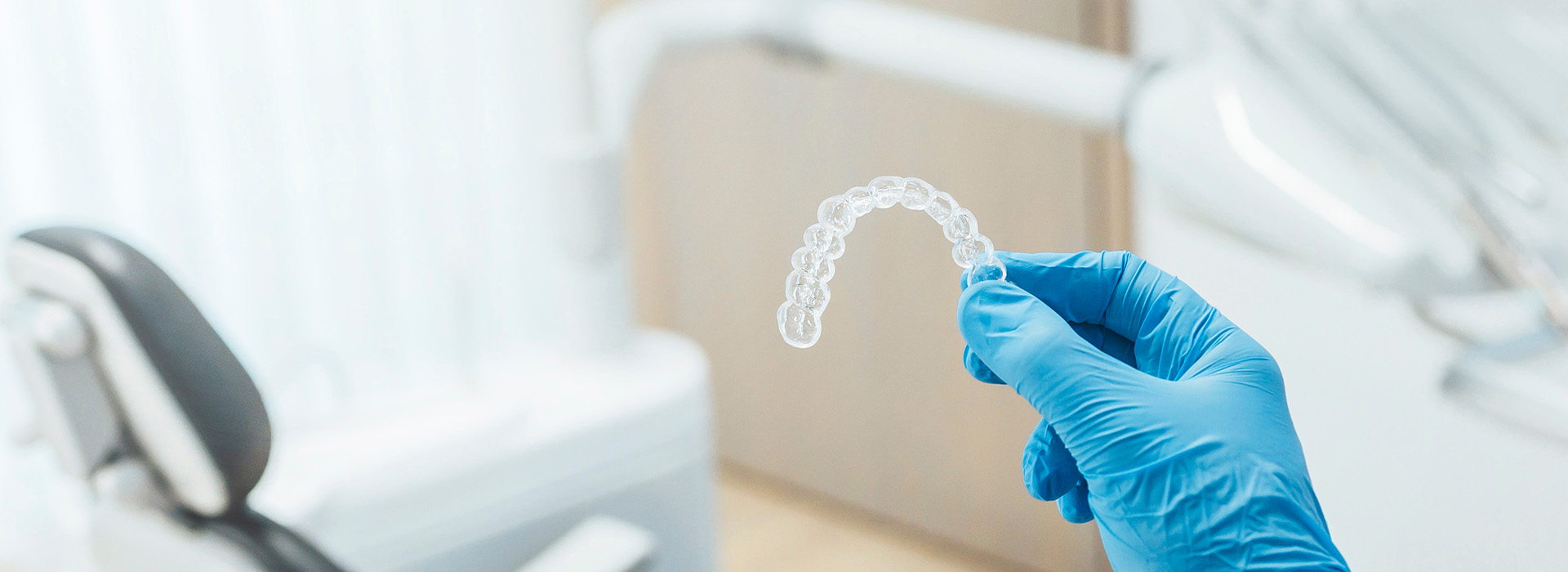 A dental office scene with a focus on a transparent mouthguard being held by a gloved hand, set against a backdrop of medical equipment and a professional environment.