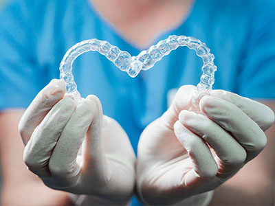 The image shows a pair of clear dental braces held by gloved hands, forming a heart shape.