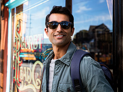 A man in sunglasses and a backpack stands outside a storefront, smiling at the camera.