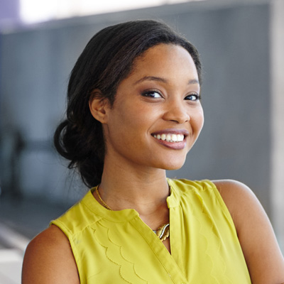 The image is a portrait of a smiling woman with dark hair, wearing a yellow top and a necklace, posing against a blurred background.