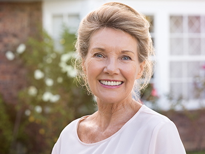 A smiling woman with short hair, wearing a light-colored top, stands in front of a house.