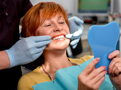 An elderly woman is seated in a dental chair, smiling at the camera, with her teeth being worked on by a dental professional.