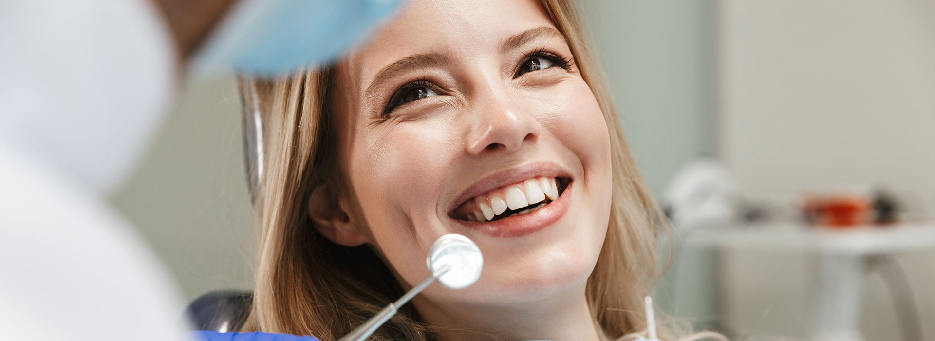 A woman is smiling at a camera while seated in front of a dental professional, who appears to be an orthodontist based on the setting and equipment.