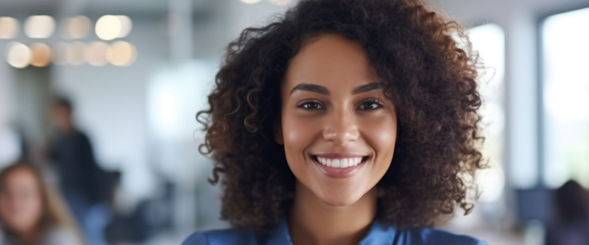 The image depicts a smiling woman with curly hair, wearing a blue shirt and standing in an office environment.