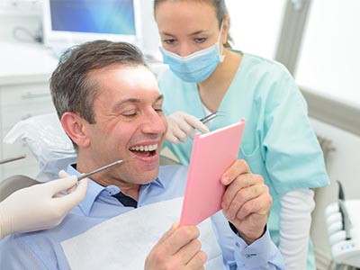 The image features a man sitting in a dental chair, holding up a pink card with a smile on his face, while a female dentist is examining him.