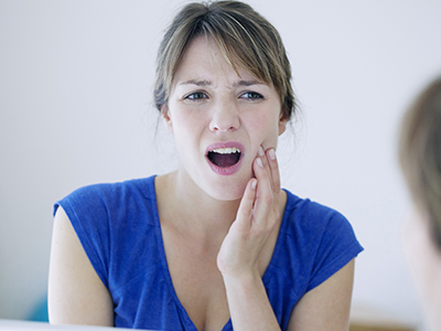 The image shows a woman with her mouth open, displaying her teeth, and appears to be in the midst of speaking or singing. She has a concerned facial expression and is looking at her reflection in the mirror.