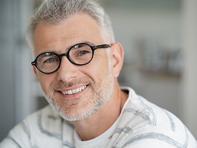 This is a photograph of an older man with gray hair, wearing glasses and a white shirt, smiling at the camera.