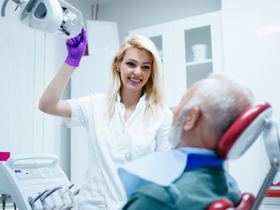 A dental hygienist is assisting a patient in a dental chair, with the patient wearing a medical face mask and receiving treatment.