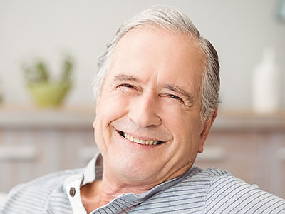 The image features an elderly man with gray hair, smiling and looking directly at the camera. He is wearing a dark shirt and appears to be seated in a comfortable setting, possibly a living room or home office.