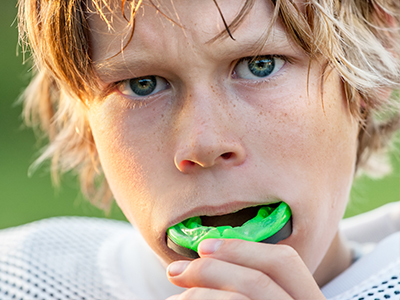 A young boy with blonde hair and a green toothbrush in his mouth, looking directly at the camera.