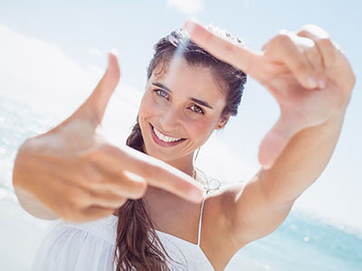 A woman with long hair is smiling and holding a camera in front of her face, capturing a selfie against a sunny beach backdrop.